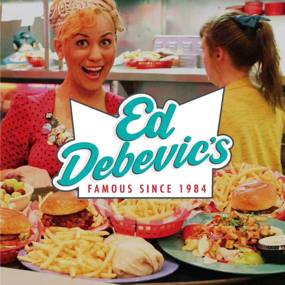 A costumed Ed Debevic’s server with a wide grin behind a counter loaded with burgers, fries and salads.