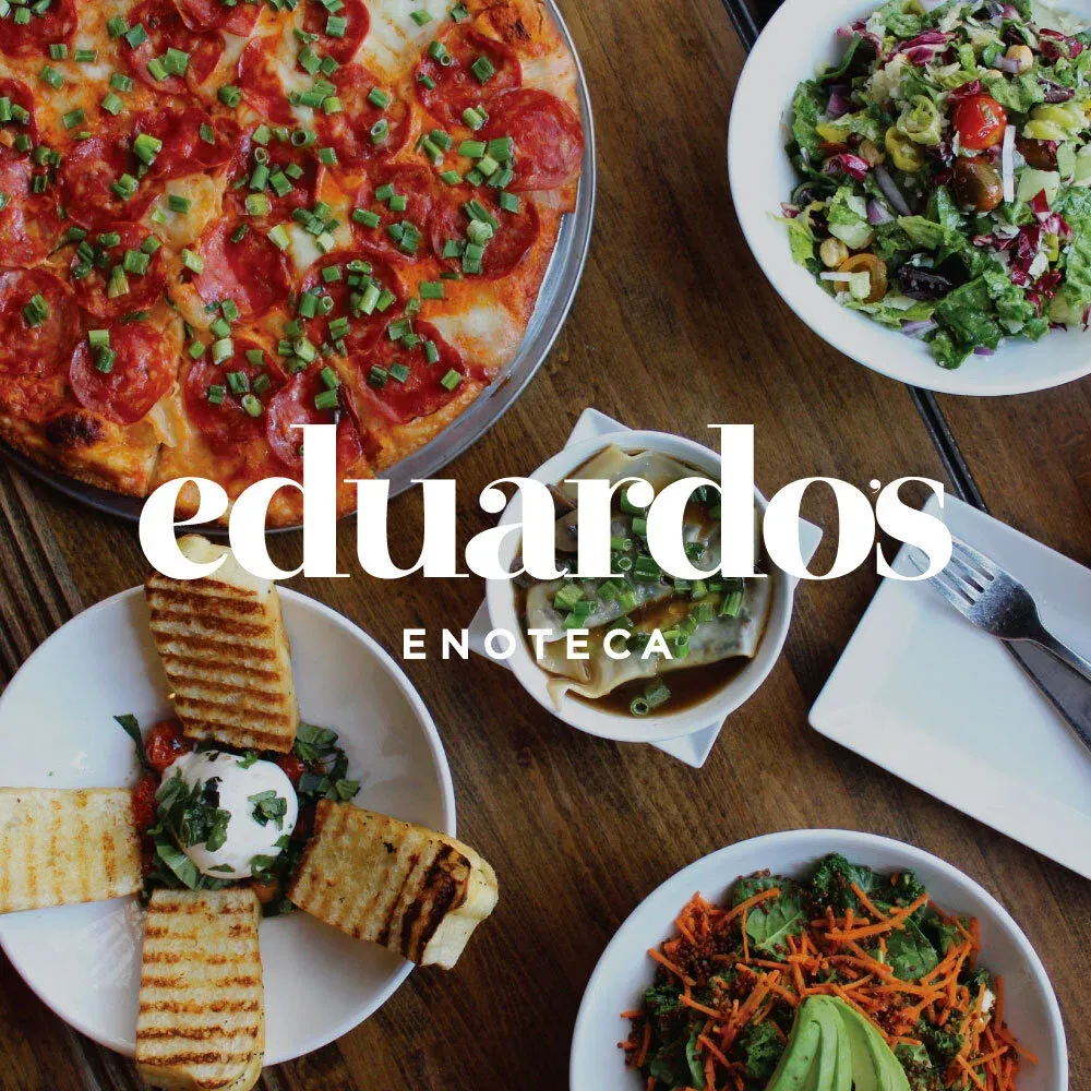 An overhead spread of Eduardo’s Enoteca dishes — pizza, charcuterie, salads and pasta — on a rustic wood table.